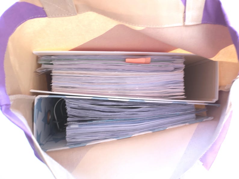 May include: A canvas tote bag with purple stripes and a beige interior holds two binders filled with papers. An orange paper clip secures the papers in the top binder. The bag is open, revealing the contents.