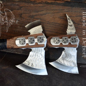 May include: Two custom-made axes with wooden handles and metal blades. The axe heads have a unique design with decorative metal accents. The handles are wrapped in black material. The image includes the text "Archangel Steel made in Detroit".