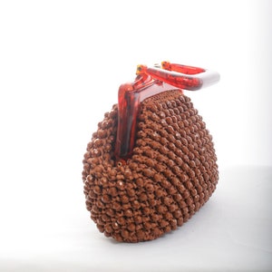 May include: A brown beaded purse with a red plastic handle. The purse is made of small, round beads that are woven together.