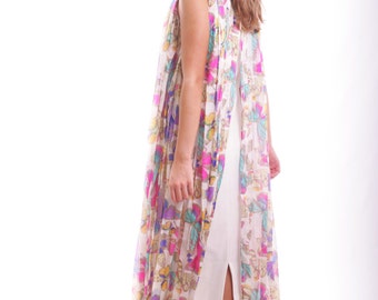 May include: A white maxi dress with a colorful butterfly print. The dress has a slit up the front and is sleeveless. The model is wearing black sunglasses.