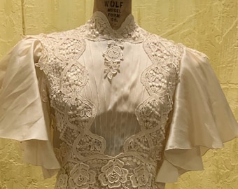 May include: A white dress with a high neckline and short sleeves. The dress is made of a sheer fabric with lace detailing. The dress is on a mannequin.
