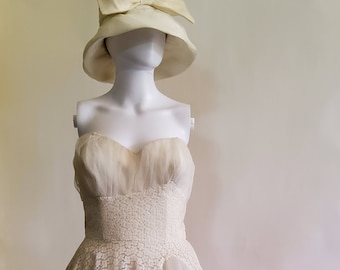 Op de afbeelding: Een witte strapless jurk met een kant overlay en een tule rok. De jurk heeft een getailleerd lijfje en een volle rok. De jurk is op een witte mannequin met een witte hoed.