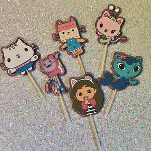 May include: A collection of six colorful character cupcake toppers. Each topper features a different cartoon cat character with glittery pink borders, attached to wooden sticks. The characters are in various poses and outfits.