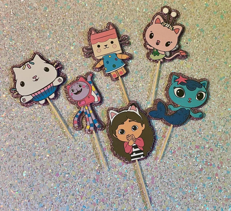 May include: A collection of six colorful character cupcake toppers. Each topper features a different cartoon cat character with glittery pink borders, attached to wooden sticks. The characters are in various poses and outfits.