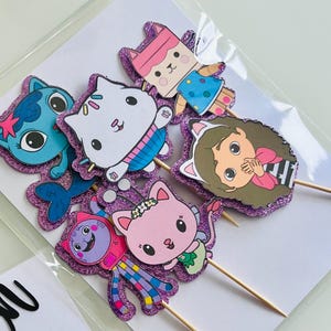 May include: A collection of colorful, glittery cupcake toppers featuring cartoon cat characters. Each topper is a different character with unique outfits and expressions, attached to wooden skewers. The toppers are set against a white background.
