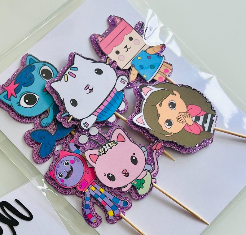 May include: A collection of colorful, glittery cupcake toppers featuring cartoon cat characters. Each topper is a different character with unique outfits and expressions, attached to wooden skewers. The toppers are set against a white background.