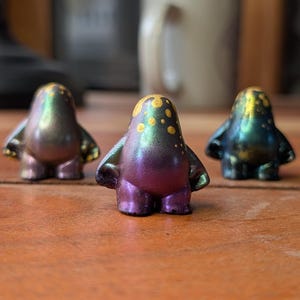 Puede incluir: Tres pequeñas figuras iridiscentes con forma de pingüino están dispuestas sobre una superficie de madera. Las figuras tienen un efecto de cambio de color, con tonos morados, verdes y dorados. Cada figura tiene pequeños puntos amarillos en la cabeza y pequeños brazos.