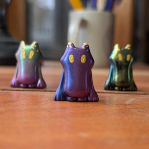 May include: Three small, cat-shaped figurines with iridescent color-shifting finishes. The figurines are purple, green, and blue, and are arranged on a wooden surface. The figurines have a stylized design with pointed ears and a simple body shape.