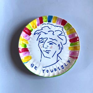 Könnte beinhalten: Ein weißer Teller mit einer blauen Strichzeichnung eines Gesichts und dem Schriftzug "BE YOURSELF". Der Tellerrand ist mit bunten vertikalen Streifen in Rot, Gelb, Grün, Rosa und Blau verziert.