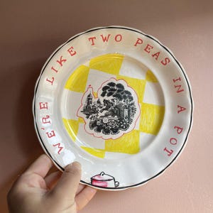 Op de afbeelding: Witte keramische plaat met een geel en wit geblokt patroon in het midden. De plaat heeft een zwarte rand en rode tekst met de tekst "WE'RE LIKE TWO PEAS IN A POT". Een zwart-wit illustratie staat in het midden.