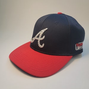 May include: A navy blue baseball cap with a red brim. The cap features a rhinestone-studded "A" logo and a small MLB team logo on the side. The cap is made of a textured fabric.
