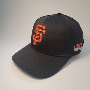 May include: Black baseball cap with an orange rhinestone San Francisco Giants logo and a Team MLB patch.