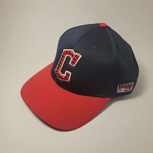 May include: A navy blue baseball cap with a red brim. The cap features a large, jeweled "C" logo in red and white. The cap has a red button on top and a "Team MLB" logo on the side.