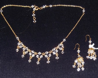 May include: A gold-toned necklace and earring set. The necklace features a delicate chain with clear crystal accents and dangling crystal pendants. The matching earrings have a similar design, with crystal clusters suspended from gold-colored hooks. The jewelry set is displayed against a dark background.