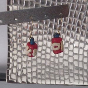 May include: A pair of red and white earrings with blue accents. The earrings are hanging from a silver metal display stand.