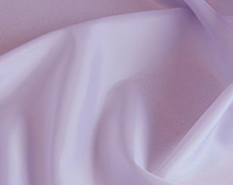 May include: A piece of light lavender fabric with a smooth, slightly reflective surface. The material is draped in soft, flowing folds that create subtle highlights and shadows across the surface, emphasizing the texture and color of the textile.