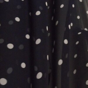 May include: Close-up of a dark navy fabric with a pattern of white polka dots. The dots vary in size and are scattered across the material. The fabric appears to be lightweight and flowing, with soft folds.