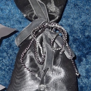 Pode incluir: Uma bolsa de cordão de cetim preto com uma fita prateada e contas prateadas. A bolsa está fechada e tem um topo franzido.