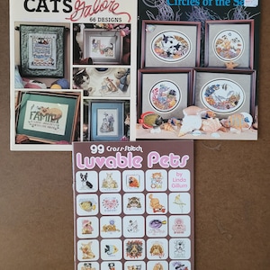May include: Three cross-stitch pattern books featuring cats, sea life, and pets. The first book is titled "Cats Galore" and features 66 designs. The second book is titled "Circles of the Sea" and features four designs. The third book is titled "Luvable Pets" and features 99 designs.