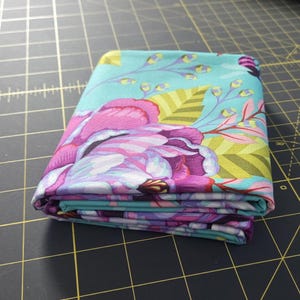 May include: A folded piece of fabric featuring a vibrant floral pattern on a light blue background. The design includes large magenta and purple flowers, lime green leaves, and thin stems with small yellow buds, all printed on a smooth textile.