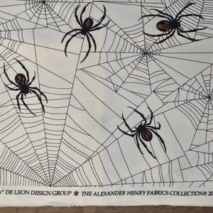 May include: Cream-coloured fabric with a repeating pattern of black spiders and spiderwebs. The spiders have brown abdomens. The text "tangled web" and "THE ALEXANDER HENRY FABRICS COLLECTION 2018" are printed at the bottom.