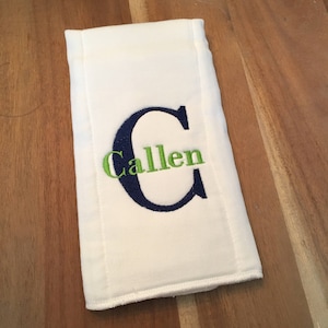 May include: White baby burp cloth with the name "Callen" embroidered in green and navy blue. The initial "C" is navy blue and the name is in green. The burp cloth is rectangular and made of a soft, absorbent material.