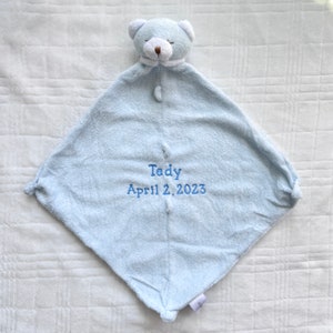 May include: A light blue baby security blanket with a teddy bear head and the embroidered text "Tedy April 2, 2023". The blanket is square-shaped with a plush texture and a button closure.
