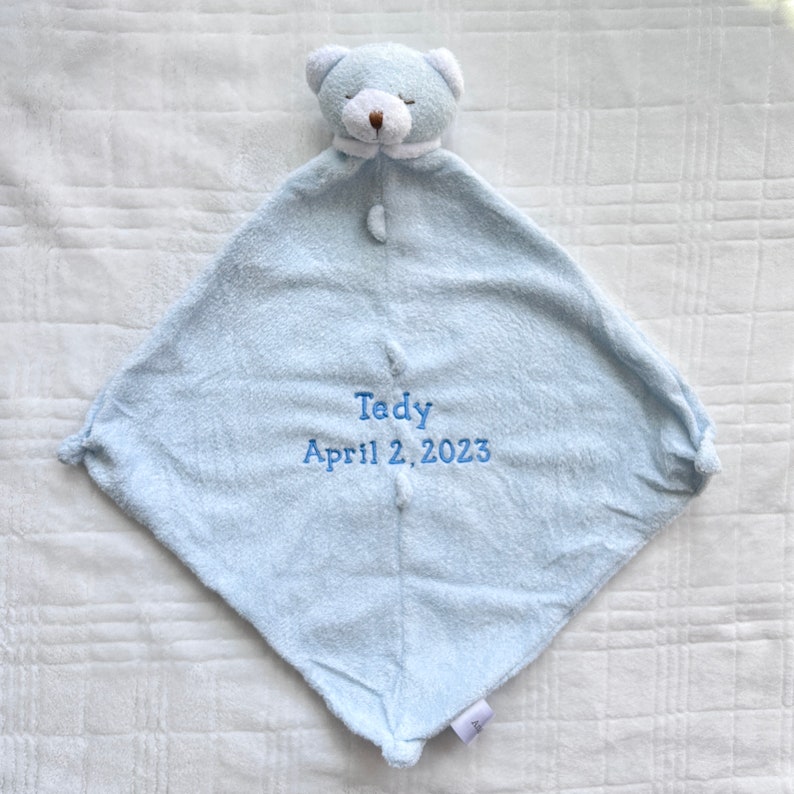 May include: A light blue baby security blanket with a teddy bear head and the embroidered text "Tedy April 2, 2023". The blanket is square-shaped with a plush texture and a button closure.