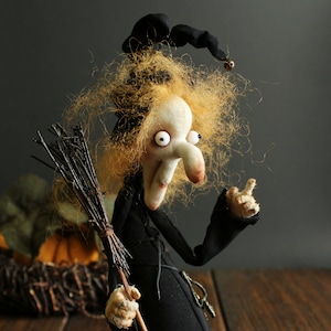 Può includere: Una bambola di strega fantasiosa con un cappello nero, capelli lunghi e biondi e un naso storto. Tiene una scopa di ramoscelli e ha grandi occhi bianchi.