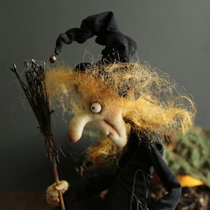 May include: A witch doll with a black hat and long, blonde hair. She is holding a broom and has a mischievous expression on her face.