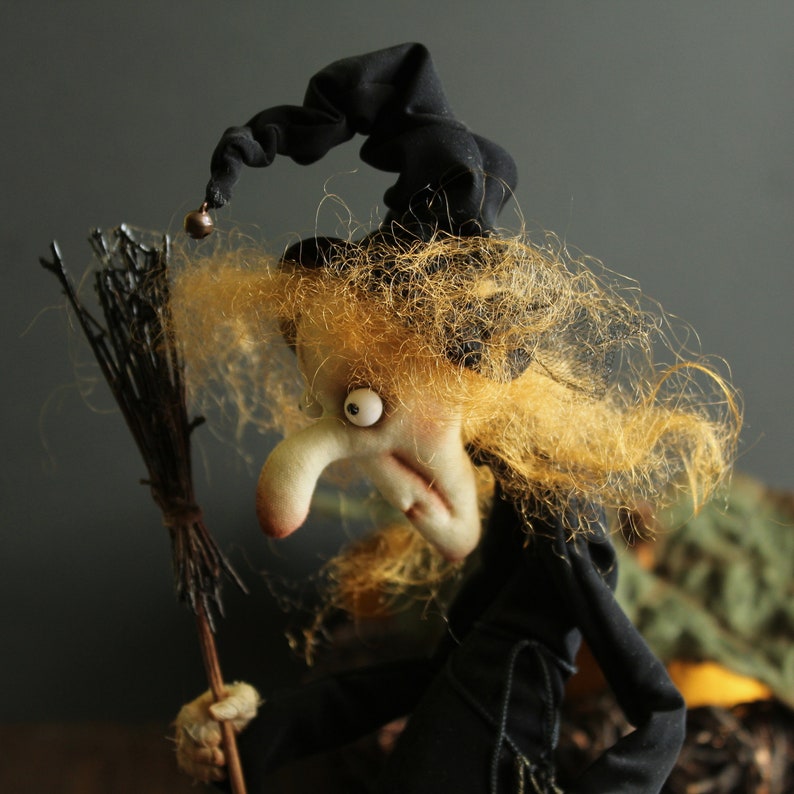 May include: A witch doll with a black hat and long, blonde hair. She is holding a broom and has a mischievous expression on her face.
