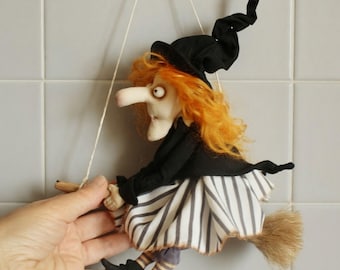 May include: A hanging fabric doll depicting a witch with bright orange hair, wearing a black pointed hat and a black jacket over a white and black striped skirt. The figure is mounted on a small wooden broomstick with a straw-like tail.