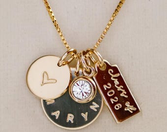 May include: Gold-toned necklace with multiple charms. Includes a round charm with a heart, a disc with the name "ARYN", a clear crystal charm, and a rectangular charm with "Class of 2026" engraved. The chain is a delicate box chain.
