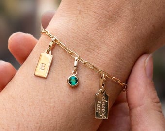 May include: A gold charm bracelet with four charms. The charms include a rectangular tag with the letter "E", a small round charm with a green stone, and a rectangular tag with the text "blessed 2021". The bracelet is worn on a wrist.