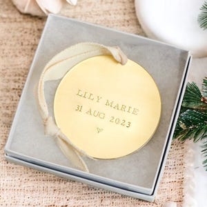 A round gold ornament with the inscription "Lily Marie 31 Aug 2023" is displayed in a gray box, surrounded by pine branches and other holiday decorations.