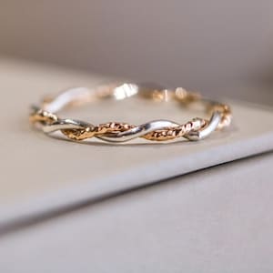 May include: A silver and rose gold braided ring. The ring features a twisted design with alternating silver and rose gold strands. The ring is displayed on a neutral-toned surface, highlighting its intricate details and metallic sheen.