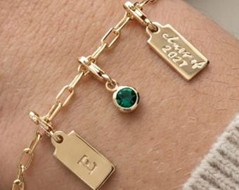 May include: Gold-toned bracelet with a chain link design. The bracelet features three rectangular charms, one with the letter "E", one with the inscription "Class of 2021", and a small green gemstone charm. The bracelet is worn on a wrist.