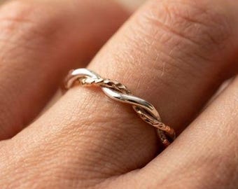 May include: A silver-toned ring with a braided design. The ring is worn on a finger, showcasing its intertwined strands. The ring's texture and design are highlighted against a soft, neutral background.