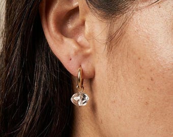 May include: A close-up of a person's ear wearing a gold hoop earring with a clear, faceted crystal pendant. The earring is dangling, and the crystal reflects light. The person has dark hair and fair skin.