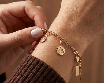 May include: Gold-colored charm bracelet featuring a chain link design. The bracelet includes several charms, such as a rainbow, a heart, a key, and a rectangular tag. The bracelet is being clasped on a wrist.