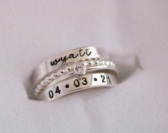 May include: Three silver rings stacked together. The top ring has the name "Wyatt" in black script. The middle ring has a small diamond. The bottom ring has the date "04 • 03 • 21".