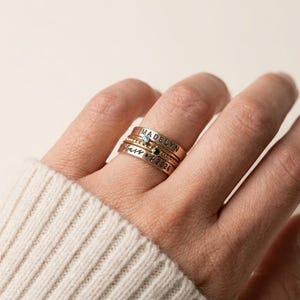 May include: A hand wearing a stack of gold rings. The rings are engraved with names and dates, and some have small gemstones. The rings are stacked on a finger, with a cream-colored sweater in the background.