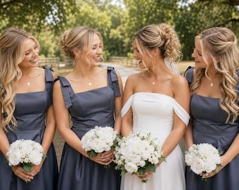 May include: Three bridesmaids in slate blue sleeveless dresses with square necklines and shoulder ties, and one bride in a white off-the-shoulder gown. Each person holds a bouquet of white roses and greenery, standing in a row outdoors.
