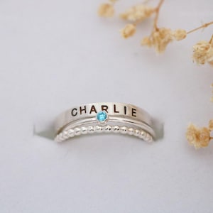 May include: A silver ring with the name "CHARLIE" engraved on the band, featuring a small, round, light blue gemstone. The ring sits atop a beaded band. Dried flowers are in the background.