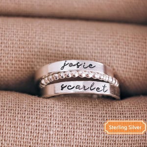A silver ring with the names "Josie" and "scarlet" engraved on it, placed on a beige fabric background.