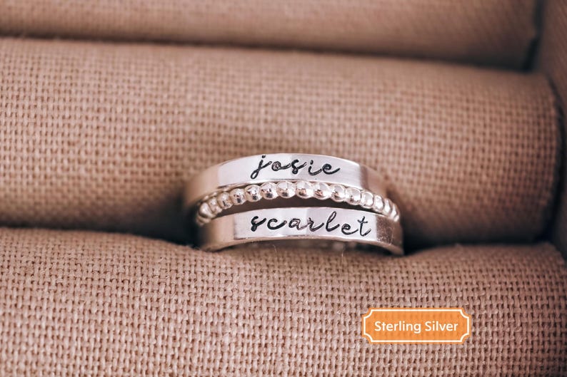 A silver ring with the names "Josie" and "scarlet" engraved on it, placed on a beige fabric background.