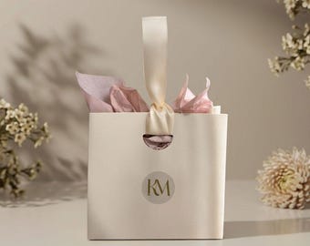 May include: A square cream-colored gift bag featuring a central circular logo with the letters KM in gold. The bag has a single cream satin ribbon handle threaded through a cutout at the top, with light pink tissue paper tucked inside.