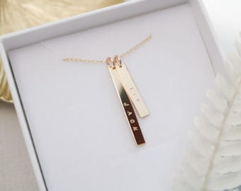 May include: A gold-colored necklace with two rectangular pendants, one engraved with "JACK" and the other with "LIV". The necklace is displayed in a white box, with a gold leaf and white fern in the background. The necklace is a personalized jewelry item.