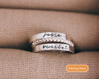 May include: Three silver rings stacked together. The top and bottom rings are engraved with the names "josie" and "scarlet" in cursive. The middle ring has a beaded design. The rings are displayed on a tan fabric surface. The image includes a "Sterling Silver" label.