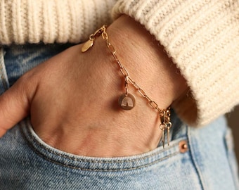 May include: Gold-colored charm bracelet with a rainbow pendant and other small charms. The bracelet is worn on a wrist, with a hand in a blue jeans pocket. The bracelet has a chain link design.